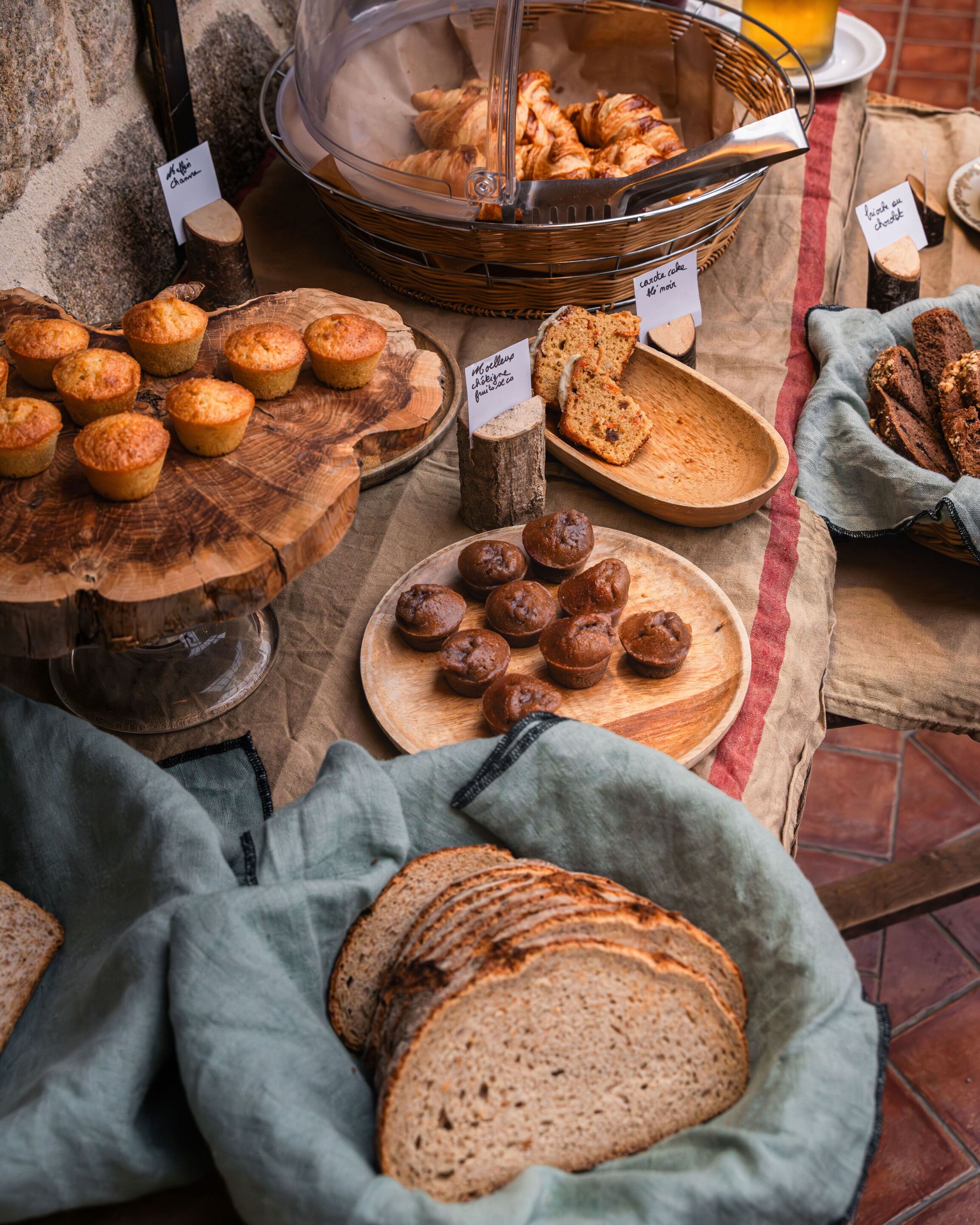 Variedad de productos horneados expuestos en tablas y cestas de madera, como muffins, croissants y pan, con etiquetas que indican los sabores.