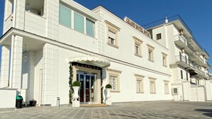 A large, white building with a grand entrance, multiple balconies, and a spacious paved area in front.