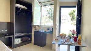 A compact hotel room with a bed, a wardrobe, a small kitchenette, and a dining area with breakfast items.