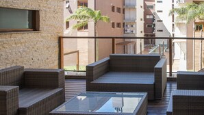 Un balcon doté de meubles en osier et d’une table en verre, donnant sur une cour intérieure agrémentée de palmiers et sur un bâtiment en arrière-plan.