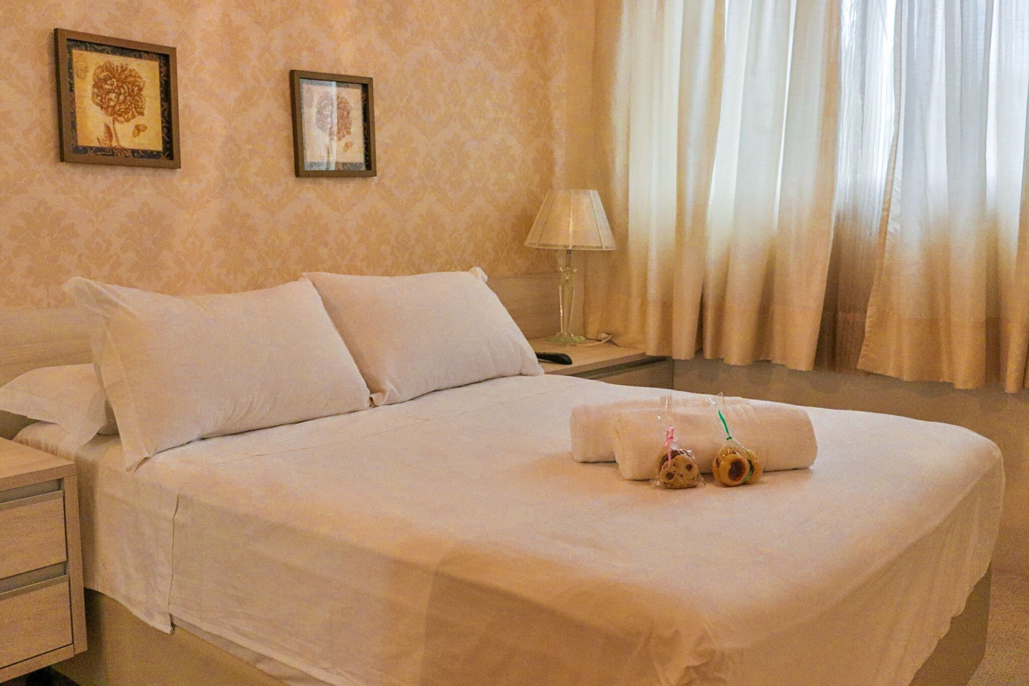Ein ordentlich gemachtes Bett mit weißer Bettwäsche, ein Nachttisch mit Lampe und zwei gerahmte Blumenbilder an der Wand.