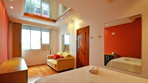 A hotel room with a bed, a sofa, a desk, a mirror, and a window. - Rio Rentals 021 - T014 Apto de 3 quarto (Rio de Janeiro)