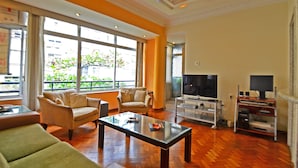 A spacious living room with a large window, a television, a glass coffee table, and a seating area with two sofas and a chair. - Rio Rentals 021 - T014 Apto de 3 quarto (Rio de Janeiro)