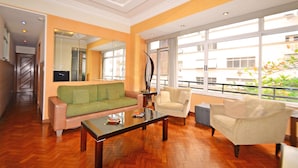 A spacious living room with a wooden floor, a green sofa, a beige armchair, and a glass coffee table. - Rio Rentals 021 - T014 Apto de 3 quarto (Rio de Janeiro)