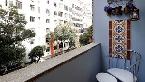 Un balcón con una silla blanca y una mesita pequeña, con vistas a una zona residencial con edificios de varios pisos y árboles.