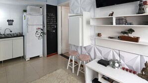 Una cocina moderna con armarios blancos, un microondas y un refrigerador. Hay una pizarra con escritura en la pared, un televisor montado sobre un estante blanco y un pequeño bonsái sobre el estante.