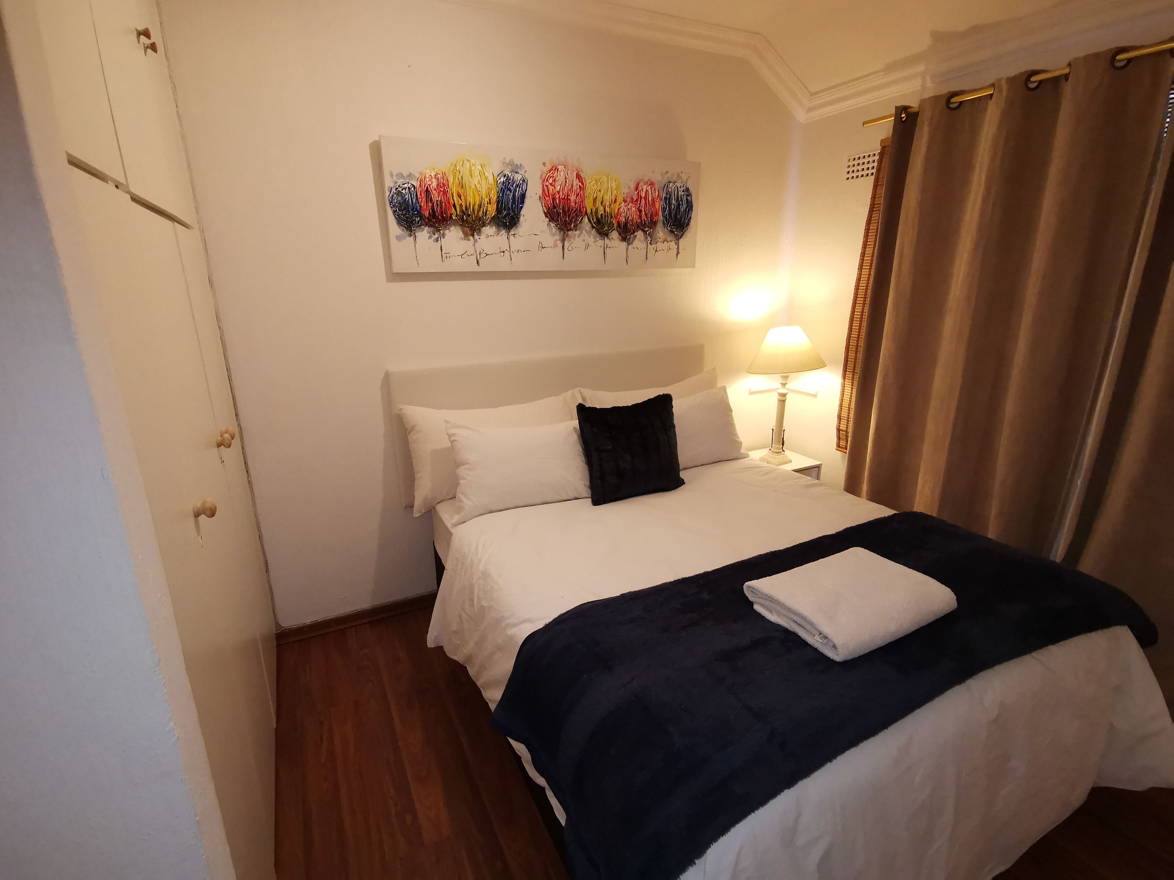 A hotel room with a bed, white walls, a painting of balloons, a nightstand with a lamp, and a window with curtains.