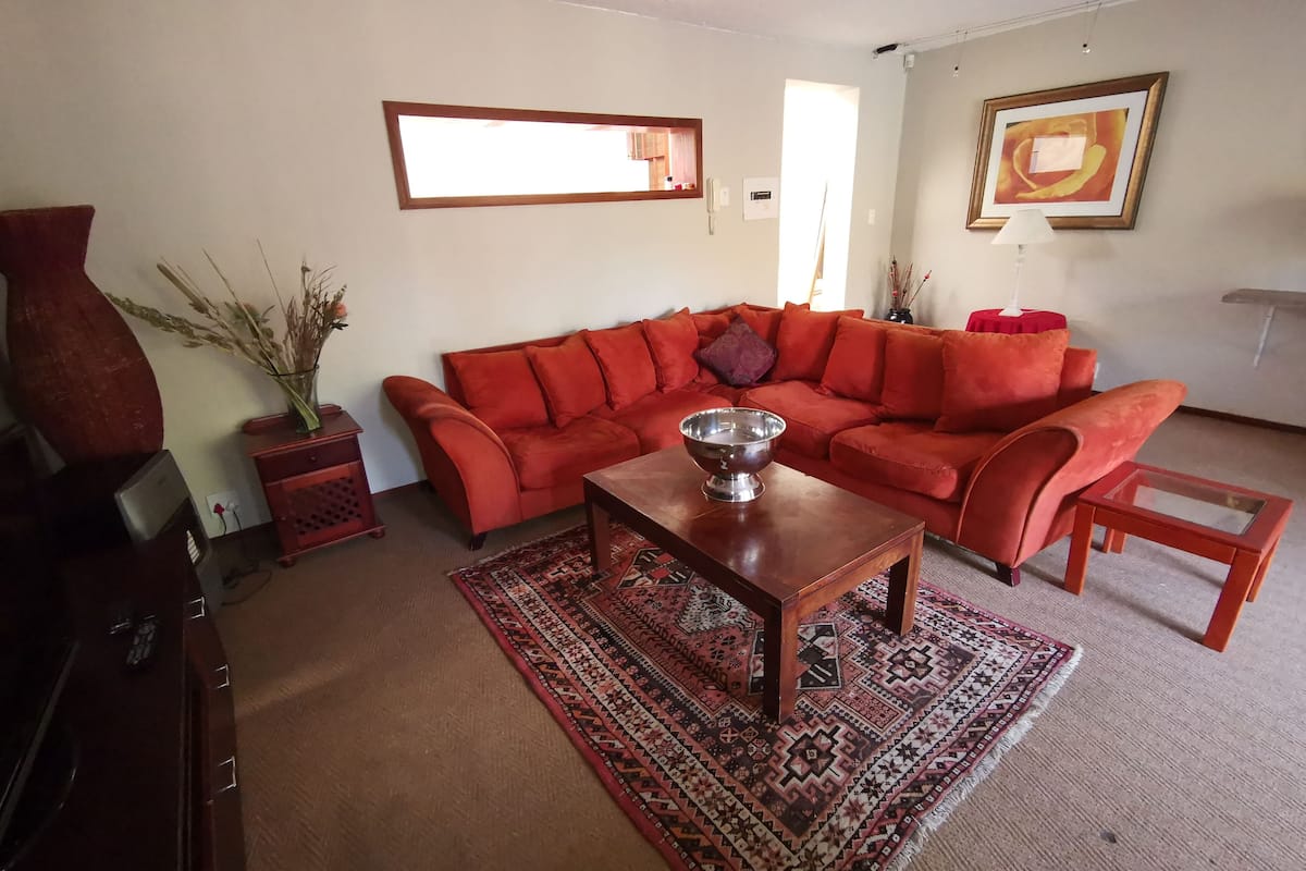 Ein Wohnzimmer mit einem roten Sofa, einem Holztisch, einem gemusterten Teppich, einer Vase mit Trockenpflanzen und einem gerahmten Bild an der Wand.