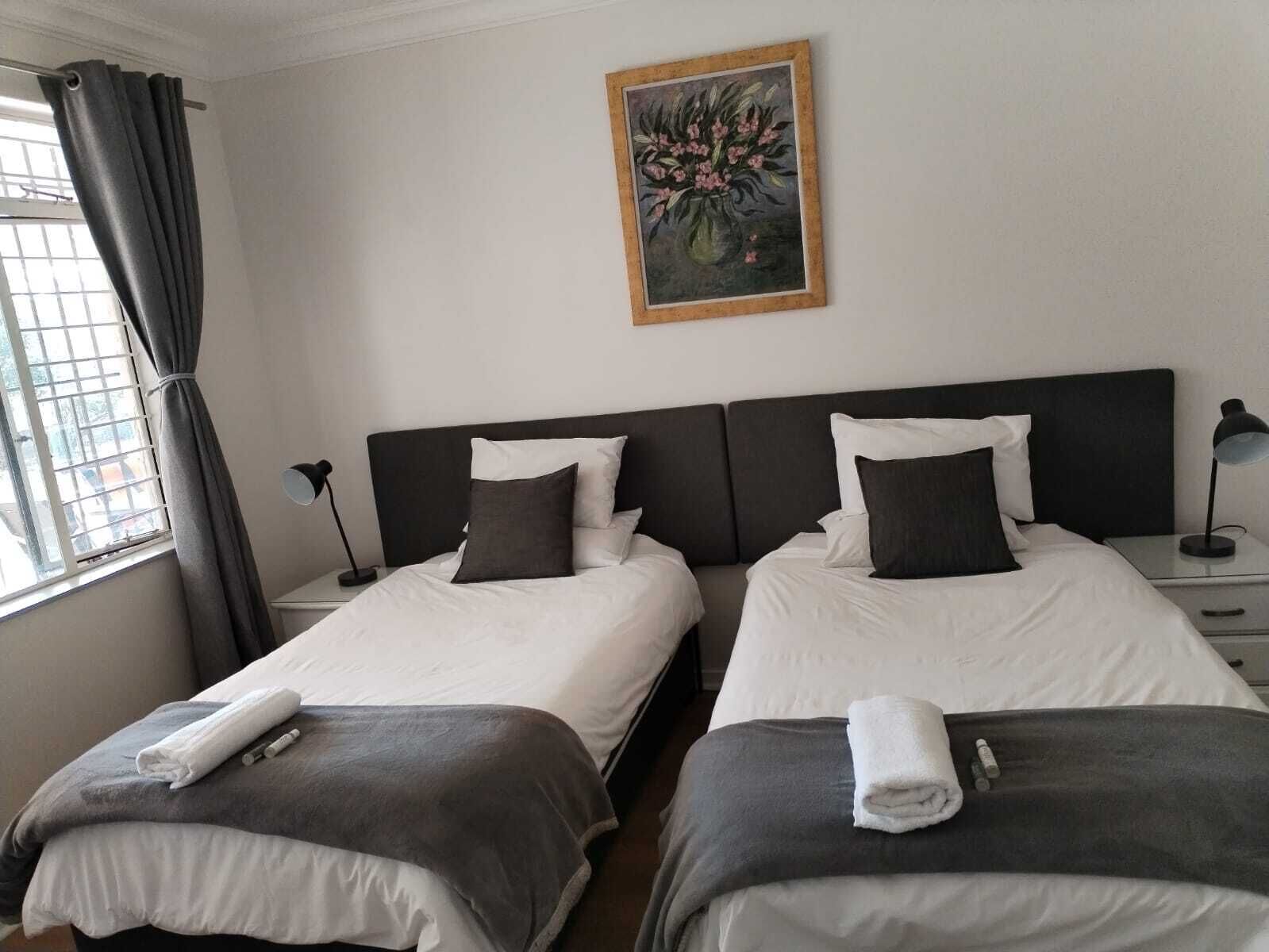 A hotel room with two beds, a painting on the wall, a window with curtains, and a bedside table with a lamp.