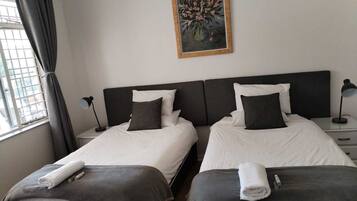A hotel room with two beds, a painting on the wall, a window with curtains, and a bedside table with a lamp.