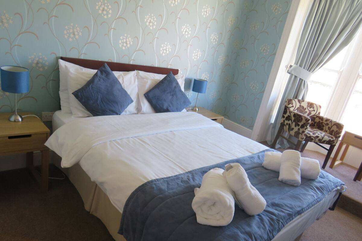 A neatly made bed with blue and white bedding, two pillows, and two rolled towels on the bed. There are two bedside tables with lamps, a chair with a floral pattern, and a window with curtains.