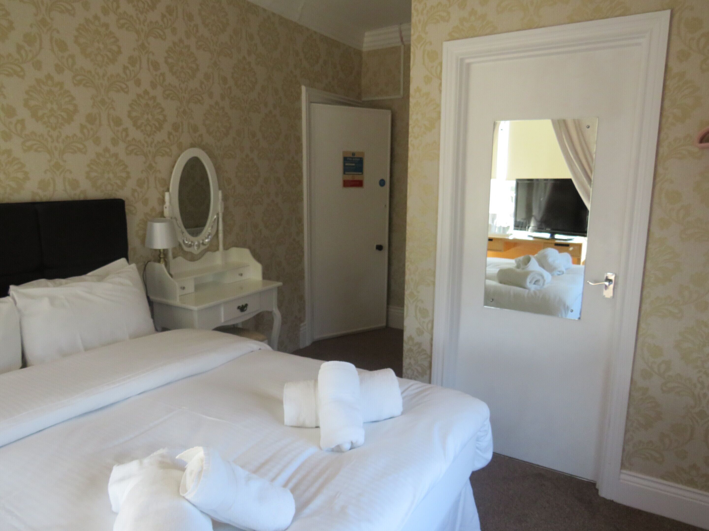 Habitación de hotel con una cama grande, una mesita de noche con espejo y una puerta que da a otra habitación.