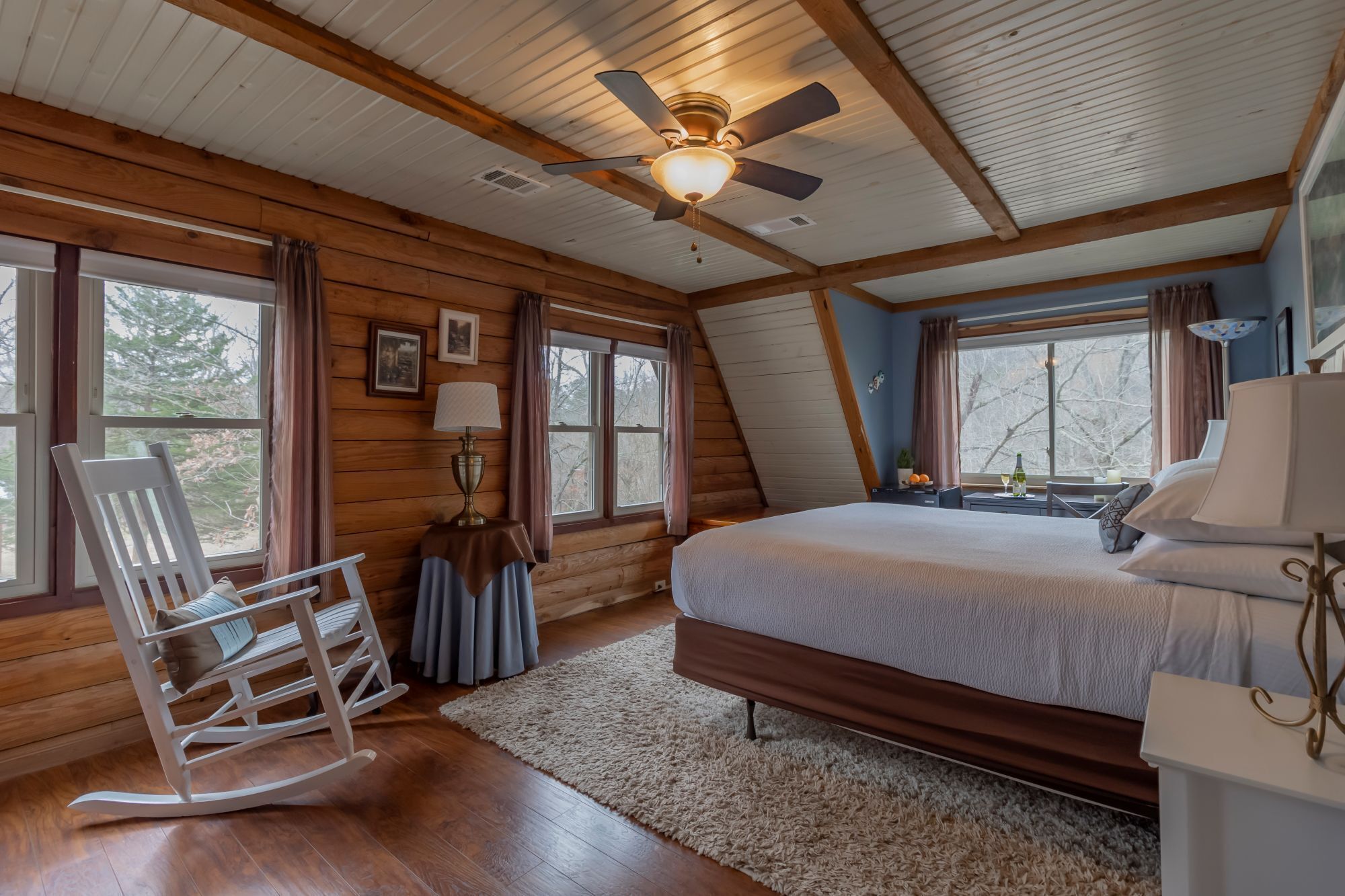 A bedroom with a bed, a rocking chair, a ceiling fan, and large windows.