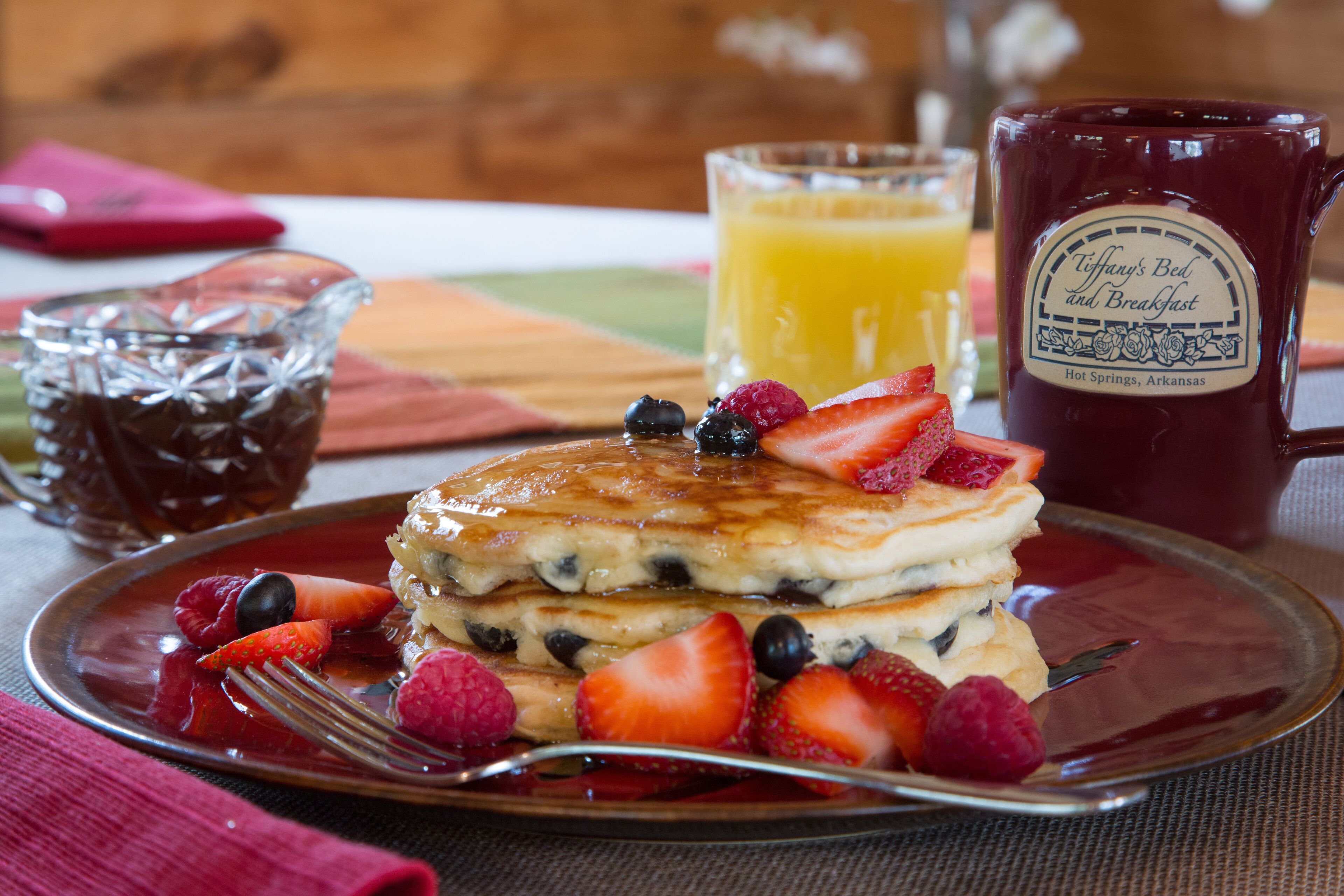 A stack of pancakes with berries and syrup on a plate, a glass of orange juice, and a mug labeled "Tiffany's Bed and Breakfast".
