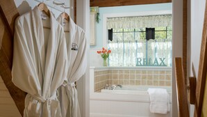 Ein Badezimmer mit einer Badewanne, einem Fenster mit blumigem Vorhang und einem Bademantel, der auf einem Kleiderständer hängt.