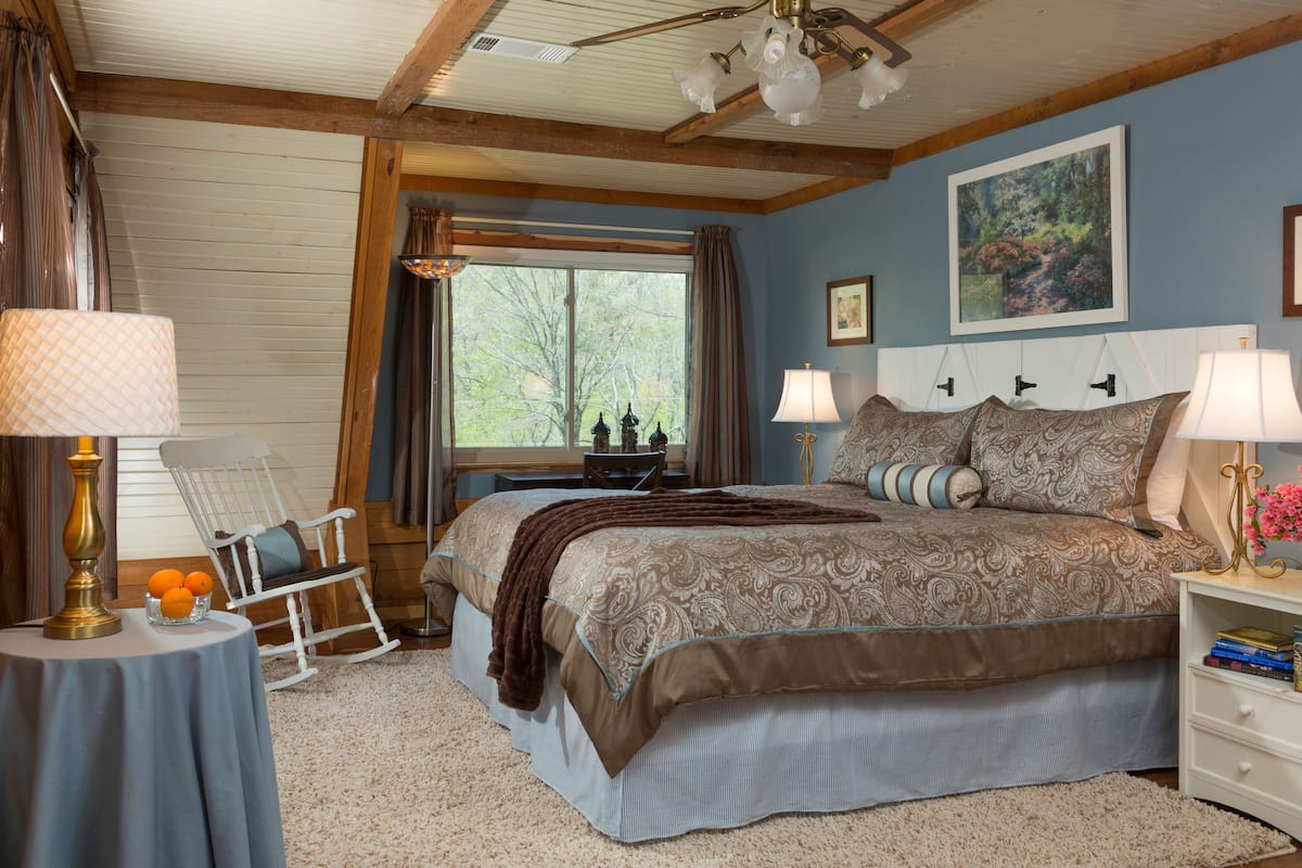 A bedroom with a large bed, two bedside lamps, a rocking chair, a small table with a lamp, and a window with curtains.