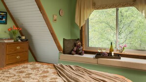 A bedroom with a sloped ceiling, a window seat with a teddy bear, a bedside table with books and a vase of flowers, and a bed with a patterned bedspread.