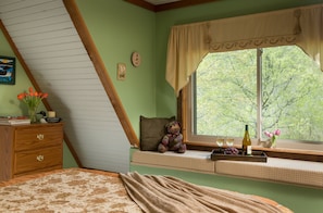 A bedroom with a sloped ceiling, a window seat with a teddy bear, a bedside table with books and a vase of flowers, and a bed with a patterned bedspread. - Tiffanys Bed & Breakfast (Bismarck)