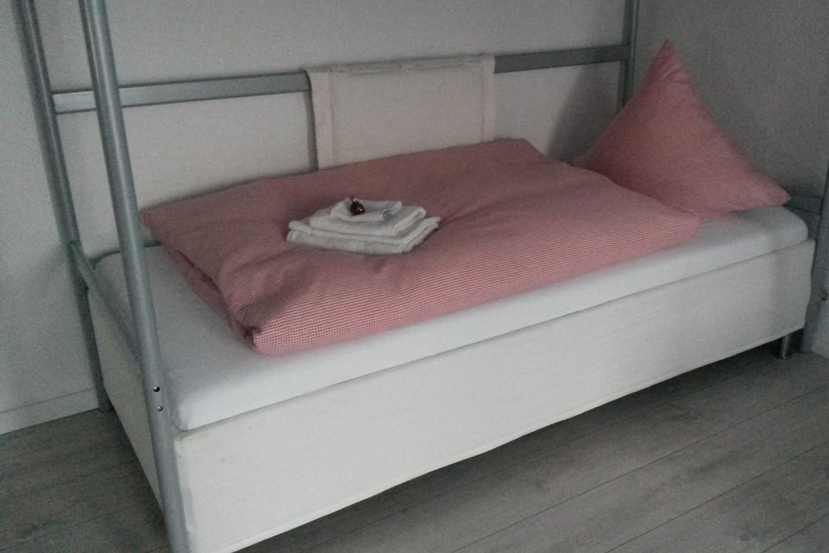 Valkoinen vuodesohva, jonka päällä on vaaleanpunainen peitto ja koristetyynyjä, taitettu pyyhe ja pieni koriste-esine.