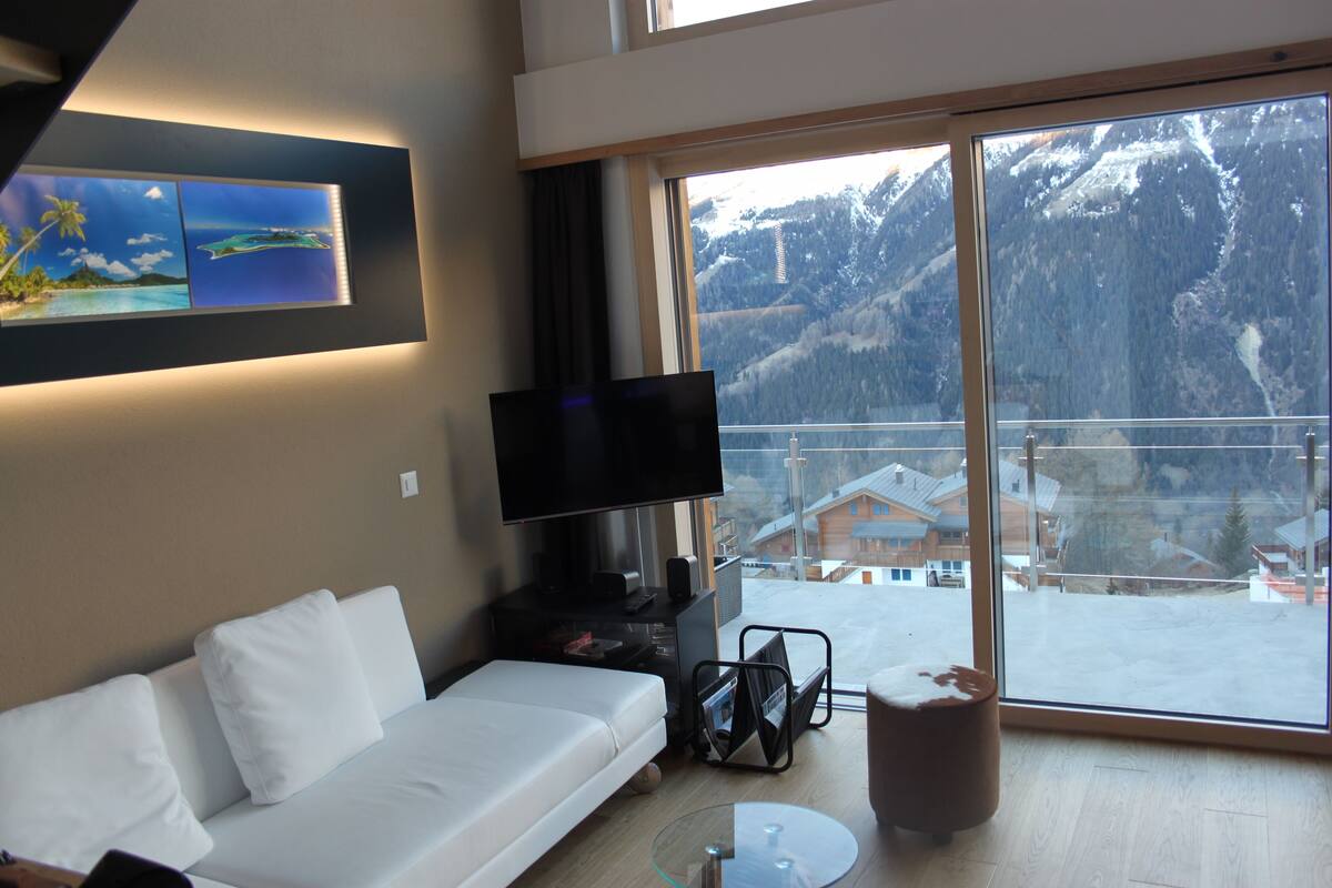 A modern hotel room with a large window offering a mountain view, a flat-screen TV, a white sofa, and a glass coffee table.