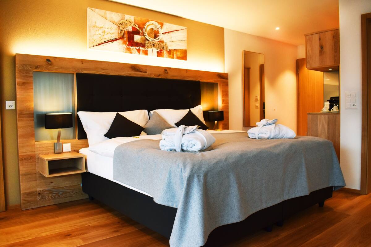 A modern hotel room with a large bed, wooden headboard, and a painting above the bed. There is a nightstand with a lamp and a small wooden cabinet.