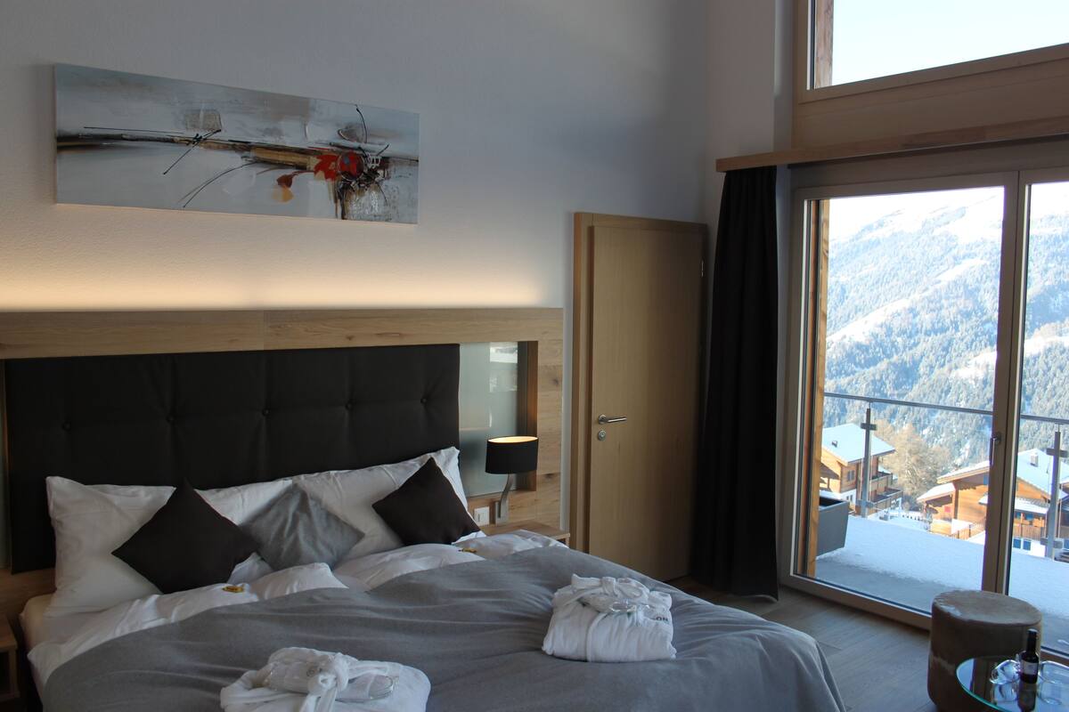 A hotel room with a large bed, a wooden headboard, a painting on the wall, and a view of a snowy mountain through the window.