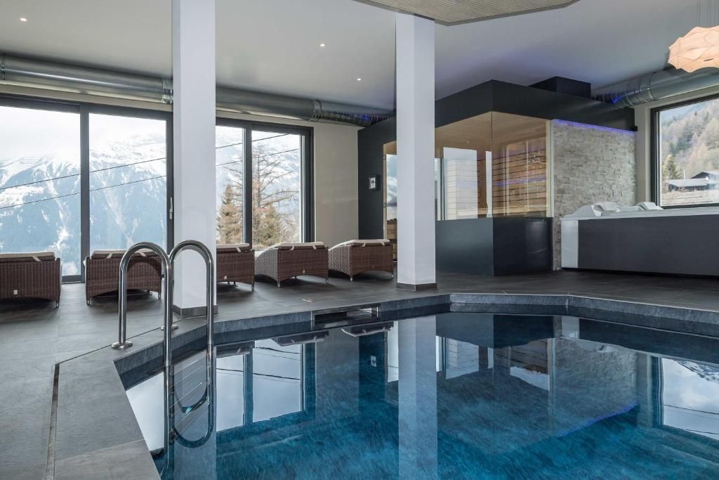 Ein moderner, überdachter Pool mit Glaswänden, Korbmöbeln und Blick auf schneebedeckte Berge.