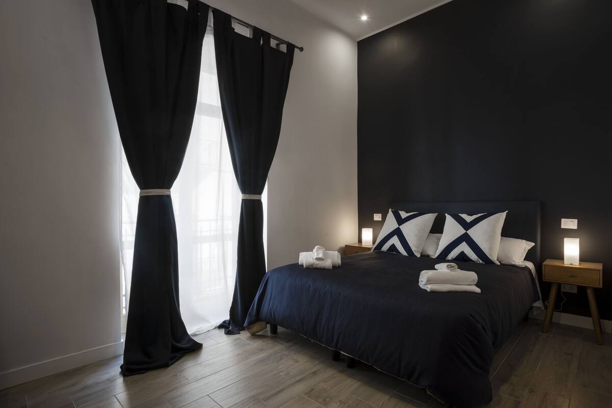 Quarto moderno com cama azul escura, almofadas brancas e mesa de cabeceira com abajur.