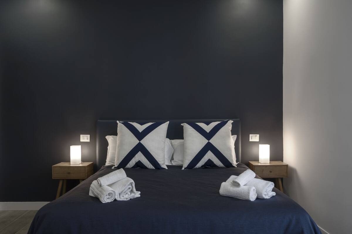 Cama bem arrumada com roupa de cama azul escuro e duas almofadas com padrão branco e azul marinho. Há duas mesas de cabeceira com luminárias e toalhas dobradas sobre a cama.