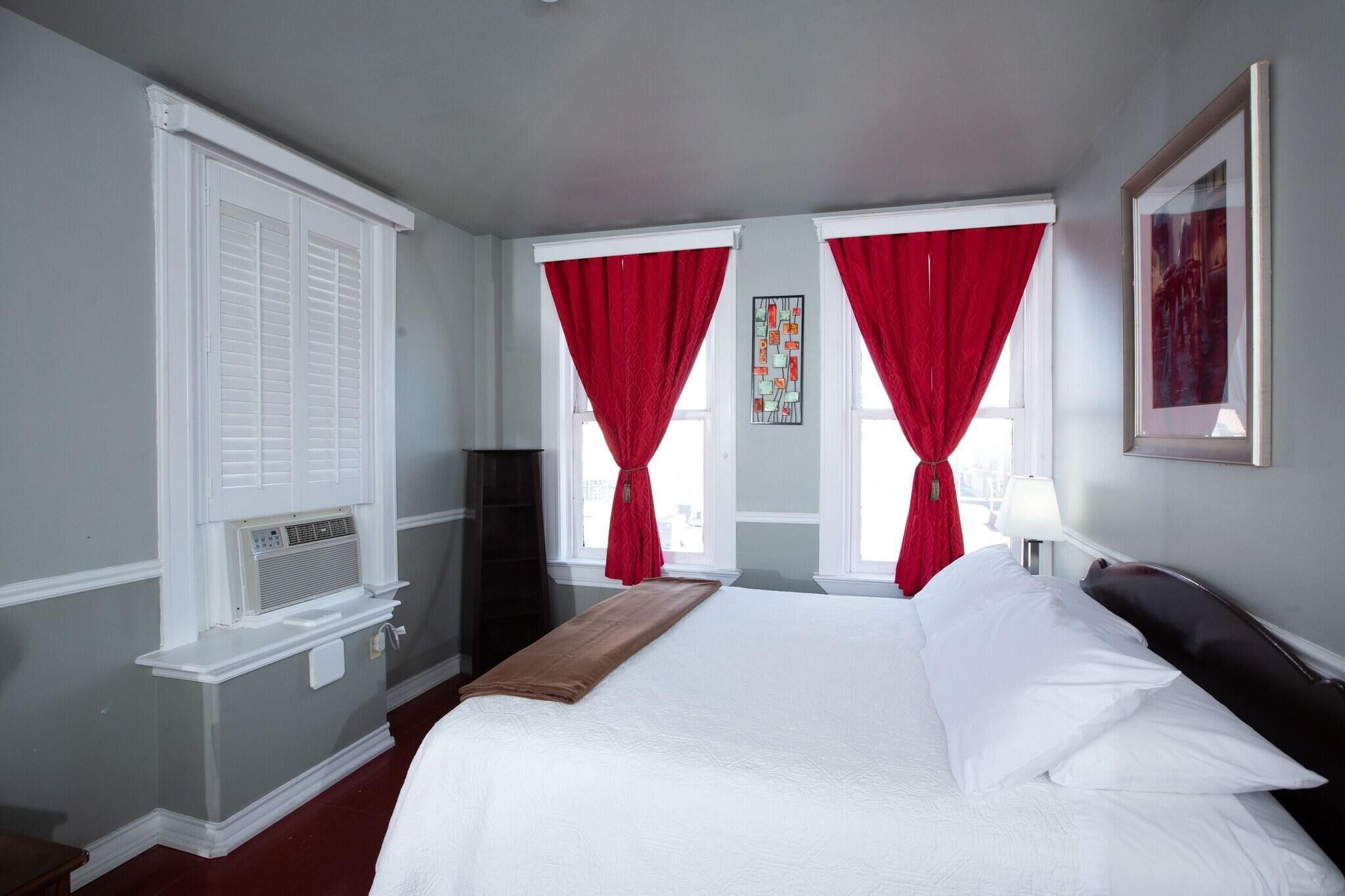 A hotel room with a bed, a window with red curtains, a radiator, and a framed picture on the wall.