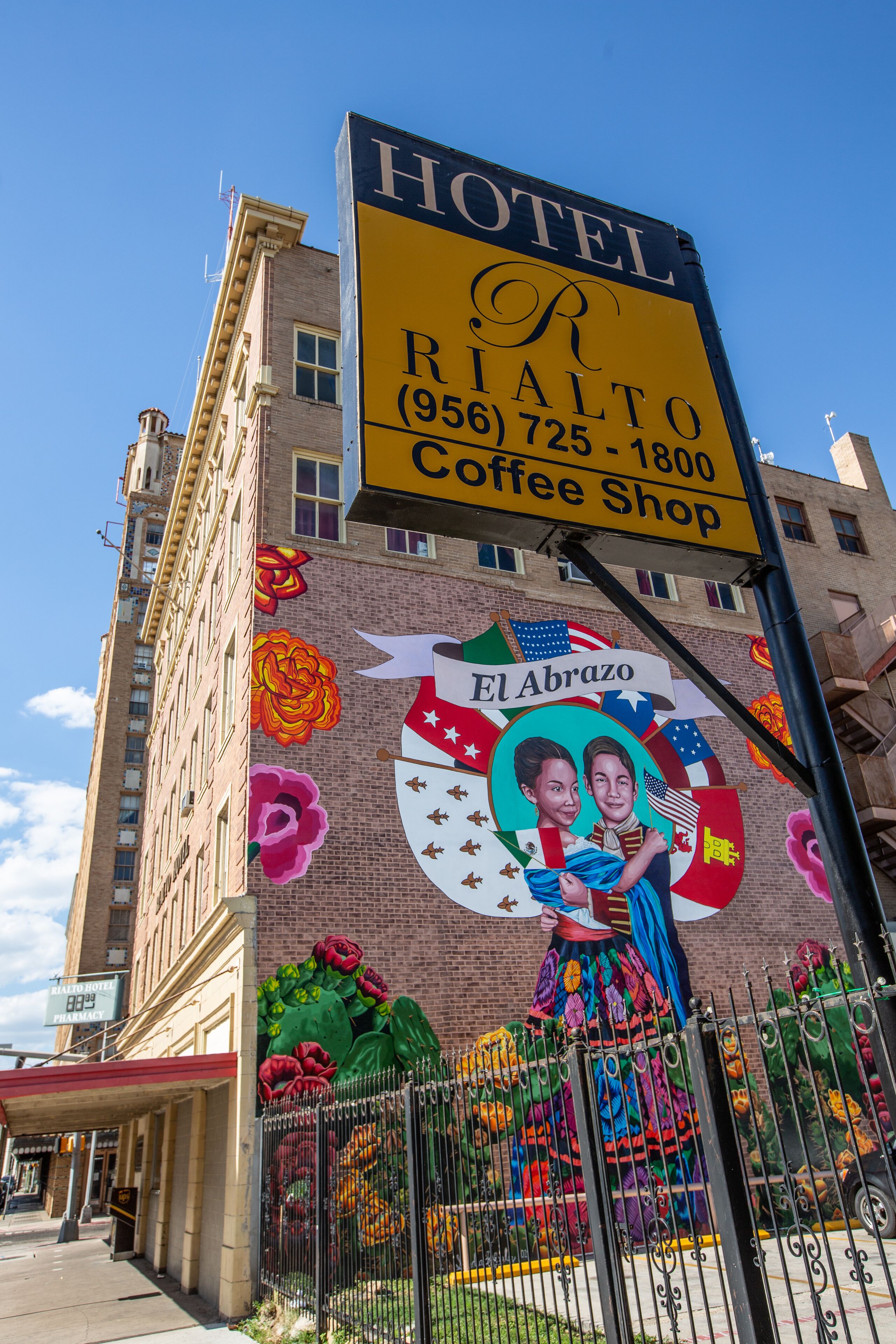 A hotel sign with a coffee shop, a mural of El Abrazo, and a brick building facade.