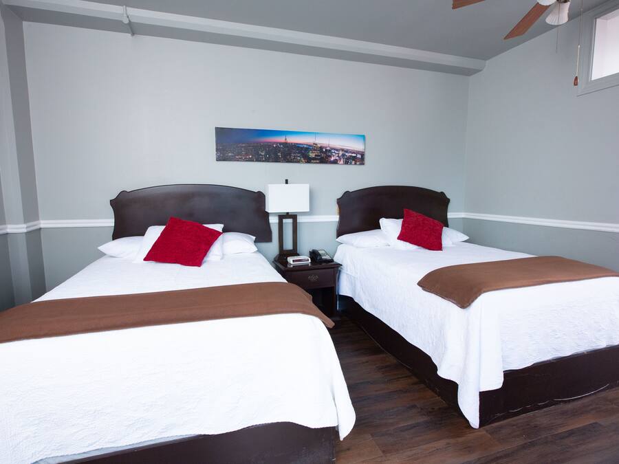 Habitación de hotel con dos camas, una mesita de noche, una lámpara y un cuadro de un paisaje urbano en la pared.