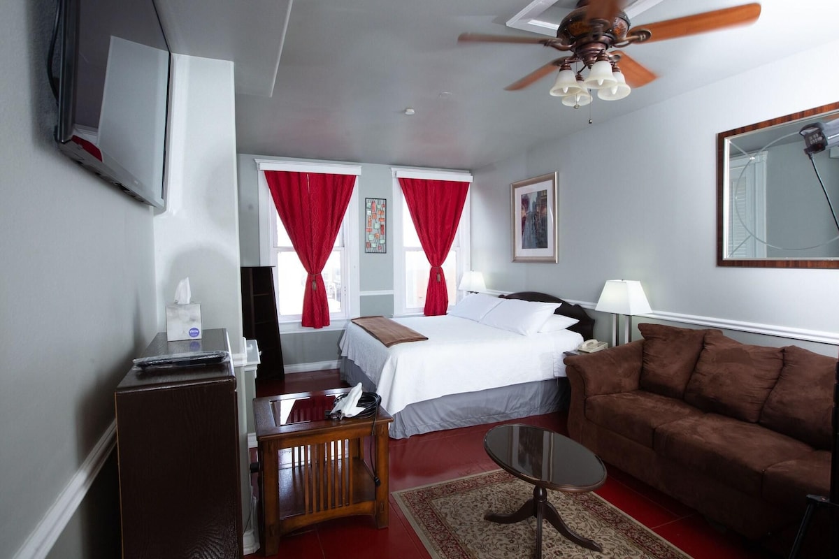 Habitación de hotel con cama, escritorio, sillón, sofá, ventilador de techo y televisión.