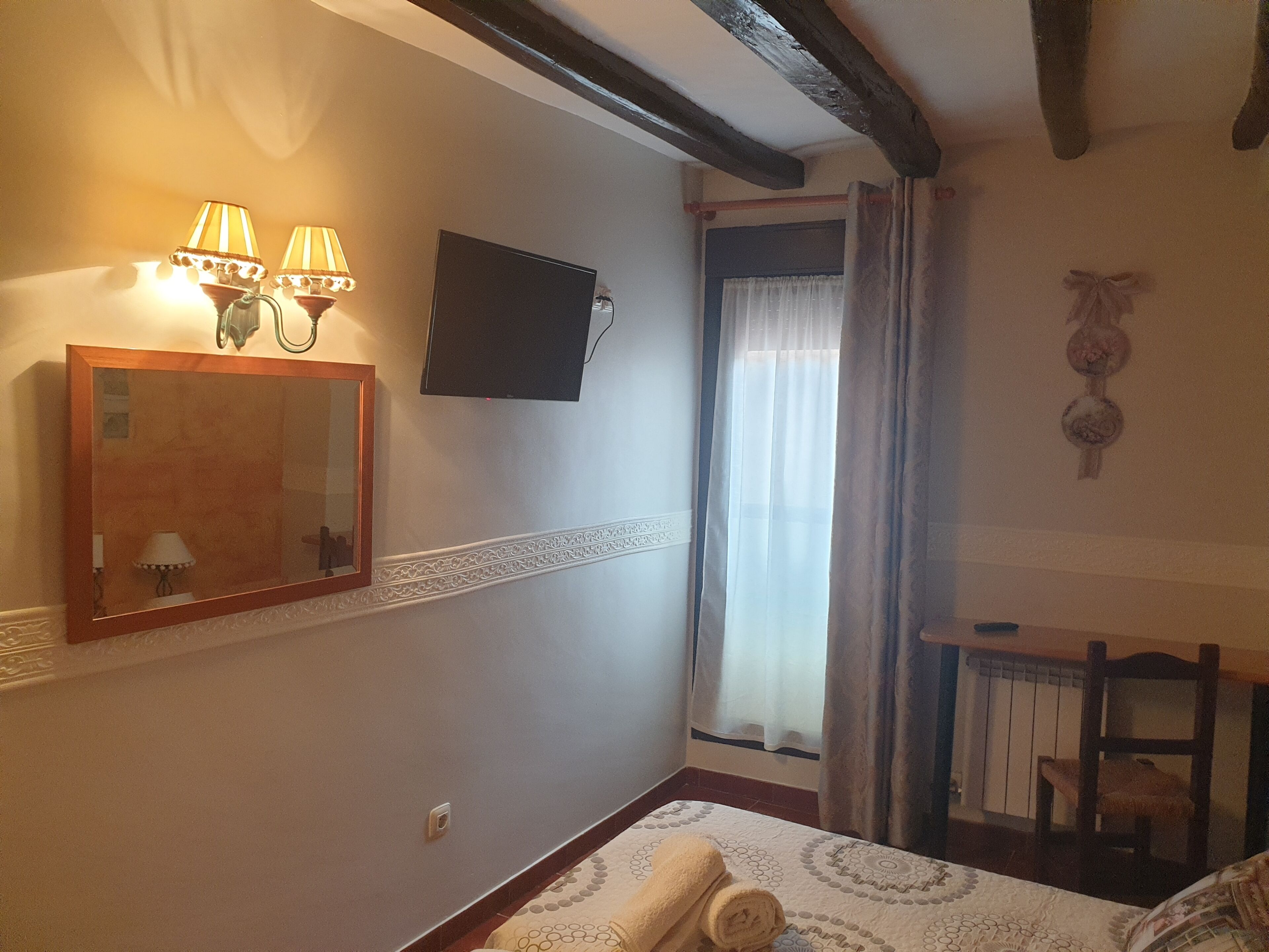A hotel room with a bed, a desk, a mirror, two wall-mounted lamps, a flat-screen TV, and a window with curtains.
