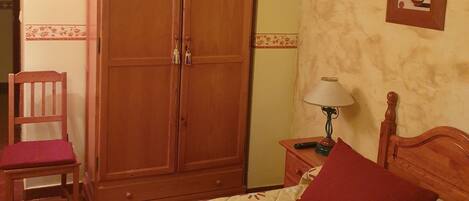 A bedroom with a bed, a wooden wardrobe, a chair, a nightstand with a lamp, and a wall-mounted picture.