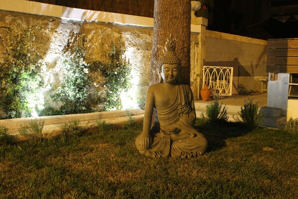 A Buddha statue in a garden setting with a tree trunk, a stone wall, and a white gate in the background. - Rosario's Boutique Apartments (Porto)