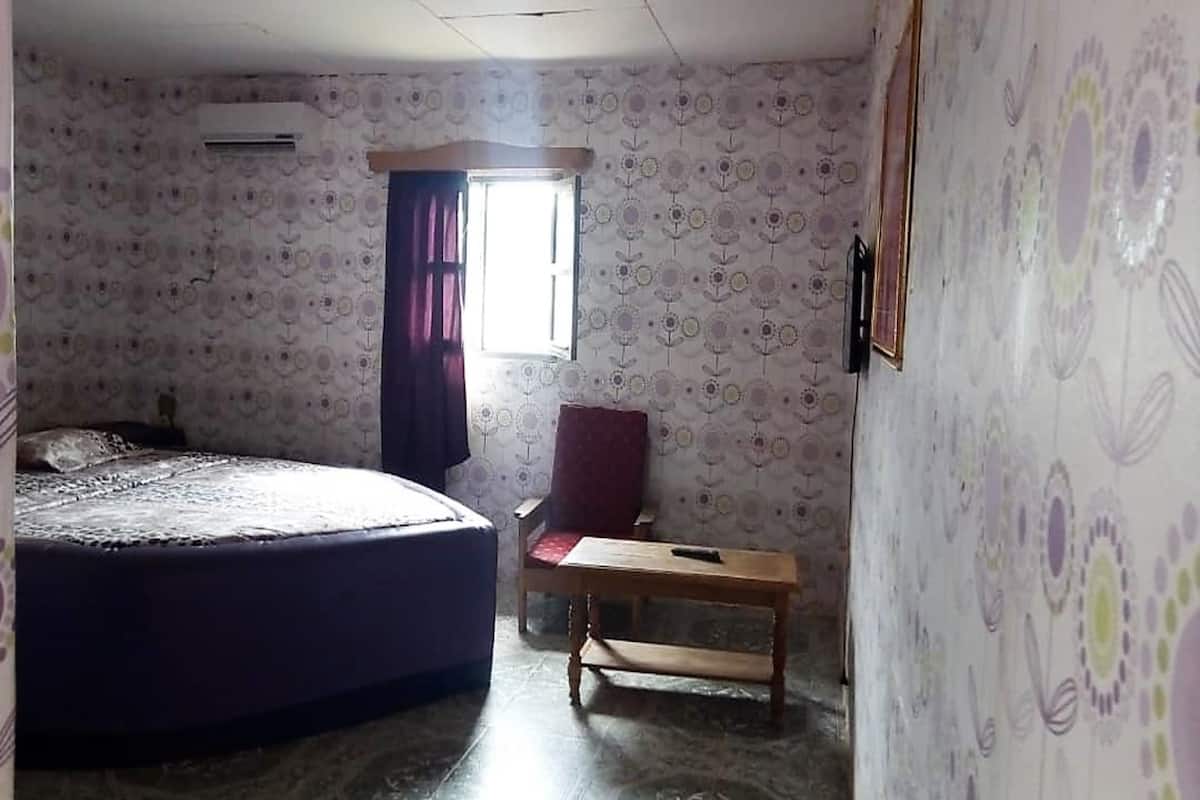 Ein Zimmer mit Bett, Stuhl, kleinem Tisch und einem Fenster mit Vorhängen.