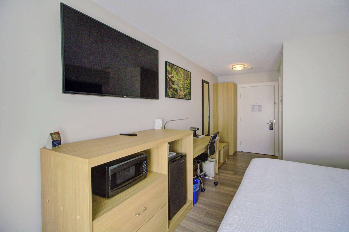 A hotel room with a bed, a desk with a microwave, a refrigerator, a TV mounted on the wall, and a door with a mirror.