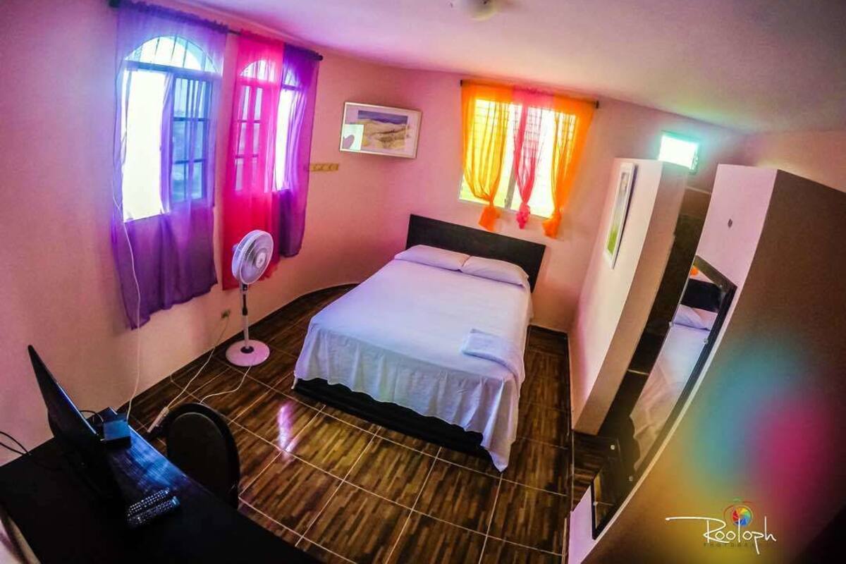 A hotel room with a bed, a desk with a computer, a fan, and curtains on the windows.