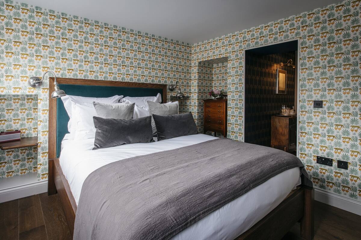 Un lit bien fait, avec une tête de lit en bois, une table de chevet avec une lampe et une commode. La pièce est décorée de papier peint à motifs et possède une porte en bois foncé.