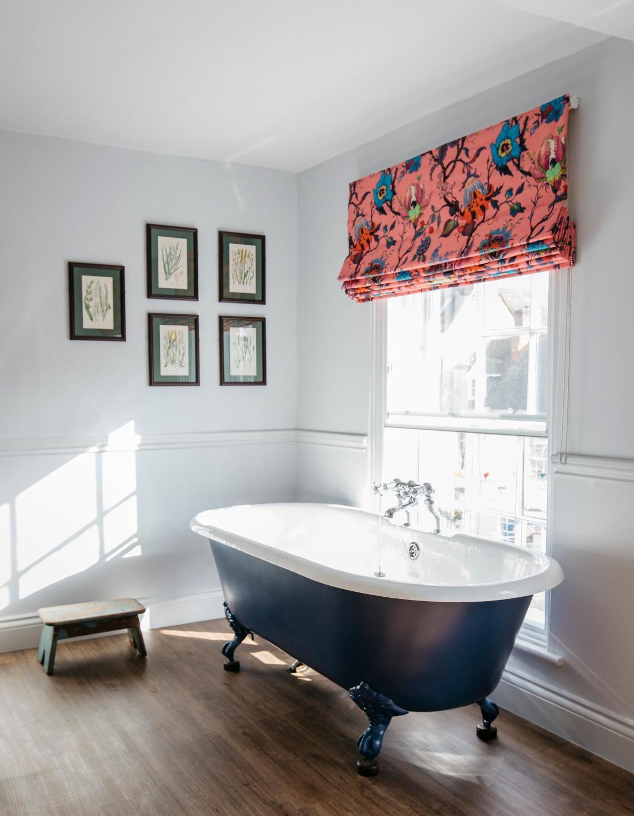A bathroom with a freestanding bathtub, framed botanical prints on the wall, and a window with a floral blind.