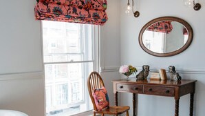 Ein Zimmer mit einem Holzbett, einem Schreibtisch mit Spiegel und einem Fenster mit Blumenrollo.