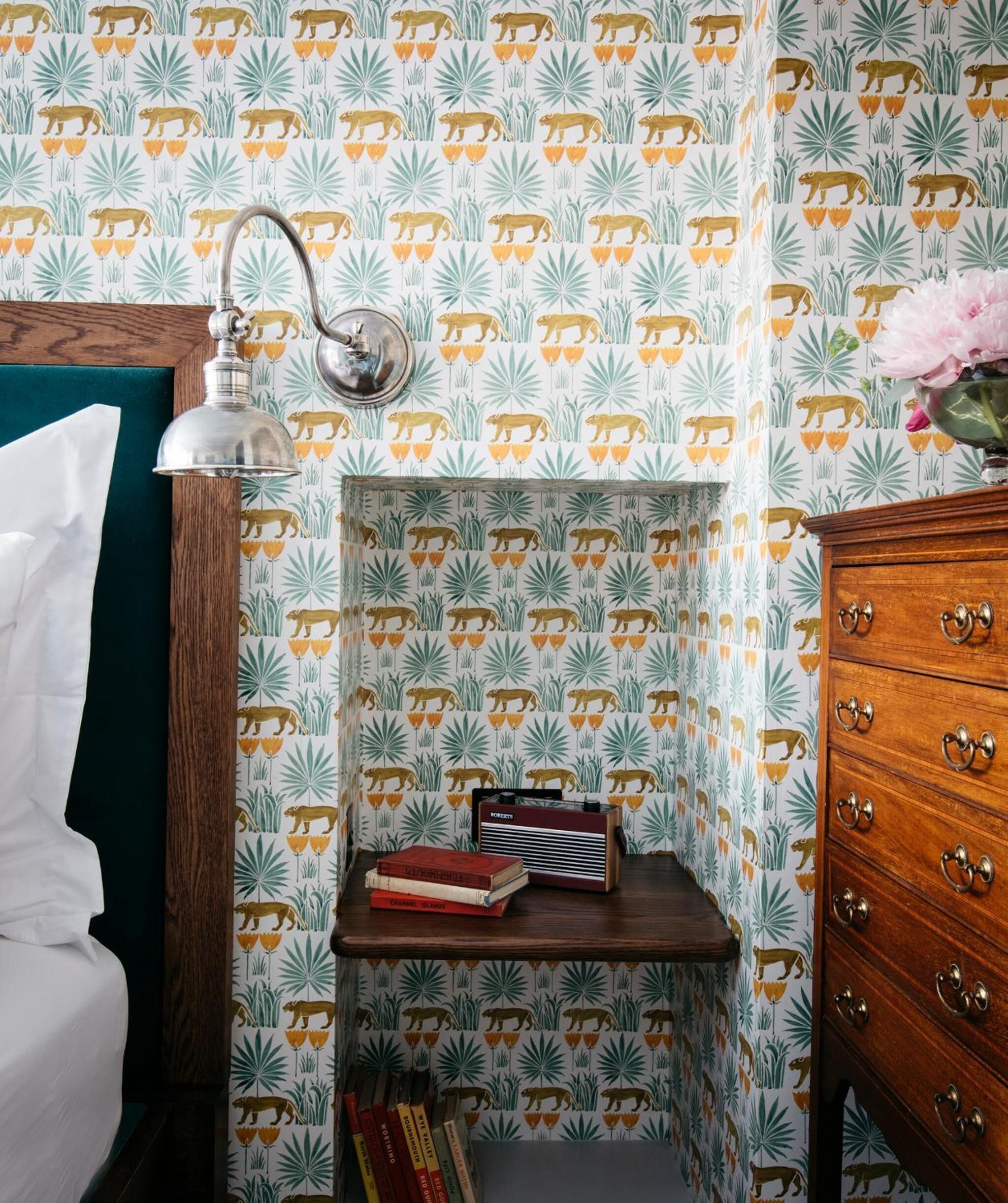 Ein Zimmer mit strukturierter Tapete mit Bären und Blättern, ein Holzschrank mit Schubladen, ein Nachttisch mit Büchern und einer Lampe sowie ein grünes Kopfteil.