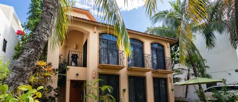 A two-story house with a balcony, a patio with a table and chairs, and a well-maintained garden with various plants and a palm tree.