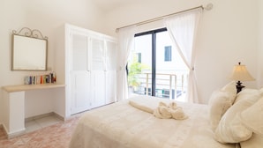 A bedroom with a bed, a bedside table with books, a window with curtains, and a view of the outside. - Condo Santuario by Playa Paradise (Playa del Carmen)