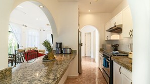 A modern kitchen with granite countertops, stainless steel appliances, and white cabinetry. There is a dining area with a red sofa and a coffee machine. - Condo Santuario by Playa Paradise (Playa del Carmen)