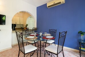 A dining area with a glass table surrounded by chairs, a kitchen in the background, and a blue wall. - Condo Santuario by Playa Paradise (Playa del Carmen)