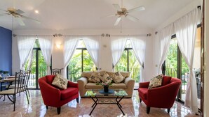 A spacious room with a beige sofa, two red armchairs, a glass coffee table, and a ceiling fan. - Condo Santuario by Playa Paradise (Playa del Carmen)