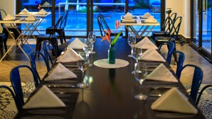 A dining area with a long table set for a meal, overhead hanging lights, and a view of a pool and lounge chairs outside. - The Blue Alcove (Da Nang)