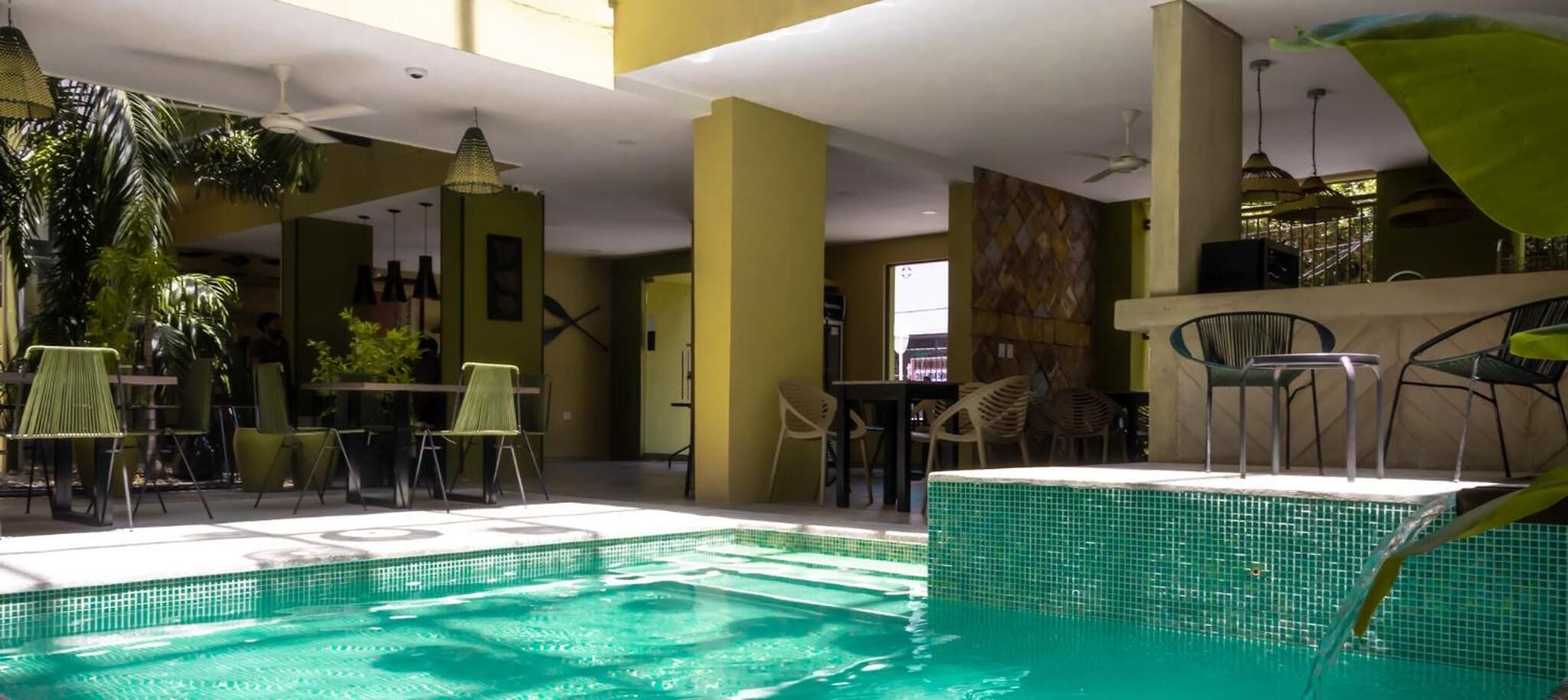 An indoor swimming pool with tiled walls, surrounded by tables and chairs, and a ceiling fan.